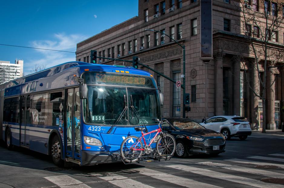 Photo of a blue bus with a bicycle in the front traveling through an intersection with a taller brown building behind it on a sunny day