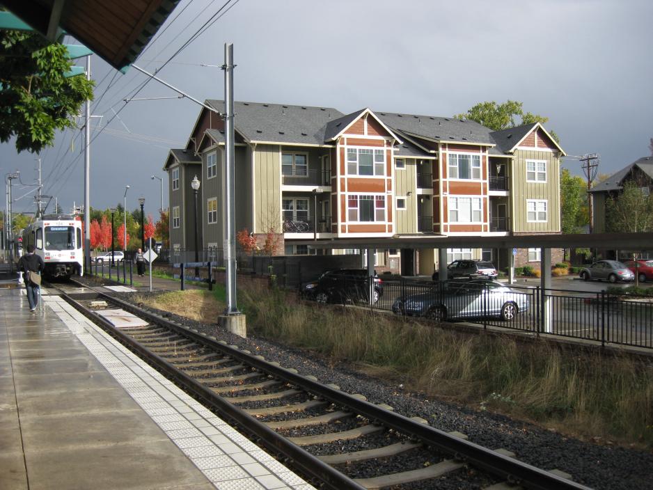 Photo of a light rail train approaching a station from the left with a person standing on the platform. There is a three-story building and parking lot in the background and it is a cloudy day