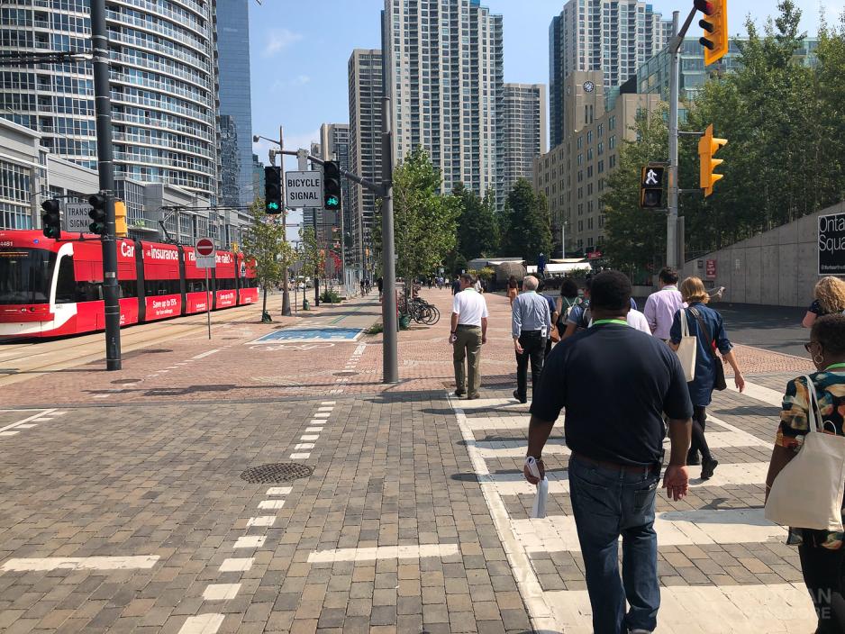 A photo of a street with people crossing the street at a pedestrian crossing, to the left is a separated bike lane, and a light rail track.