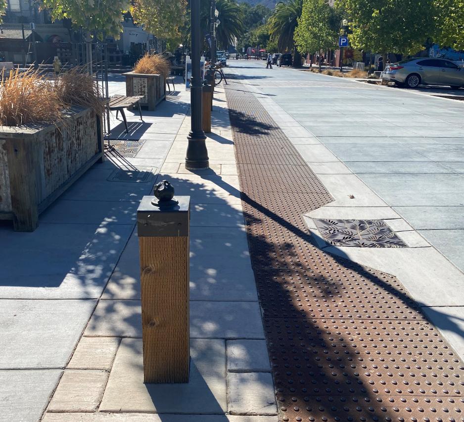 Photo of a sidewalk at the same level as the street with brown truncated domes and wooden bollards between the sidewalk space and street