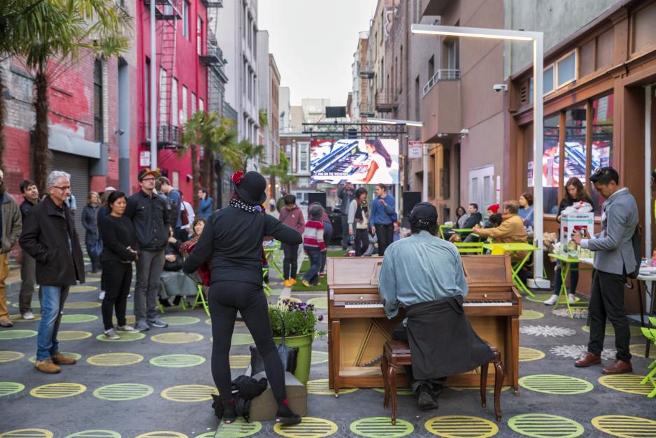 Photo of two pianists performing in the middle of a street with colorful taller buildings on both sides with polka dots painted on the ground and people watching or sitting nearby