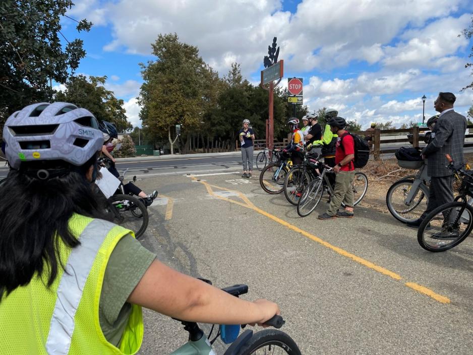woman on bike in yellow vest and helmet, other bicyclists listening to a woman speaking
