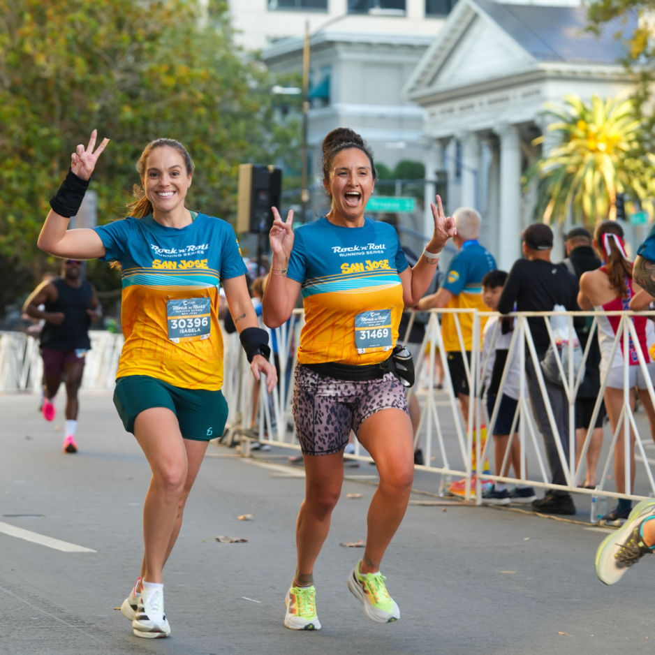 Two smiling runners wearing Rock ‘n’ Roll San José race shirts run side by side and hold up peace signs along the race course.